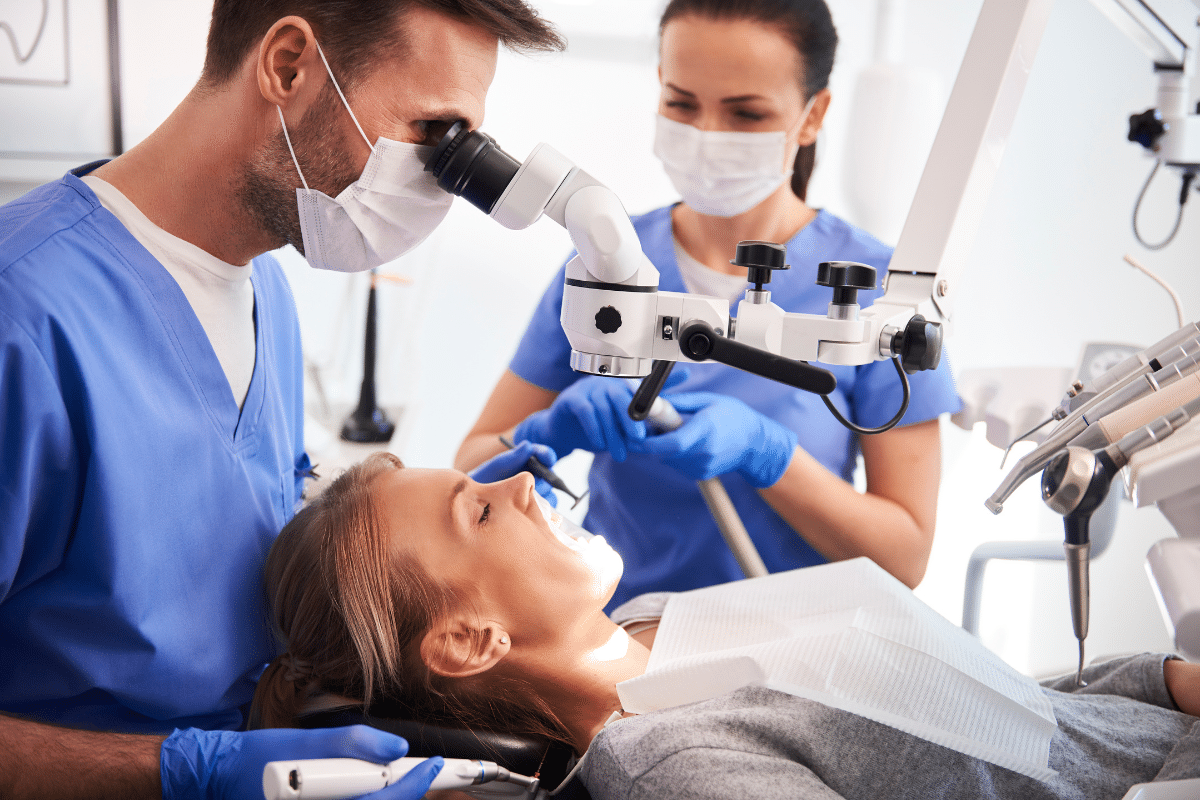 Two dentists examine young patient in dental office using microscope equipment.